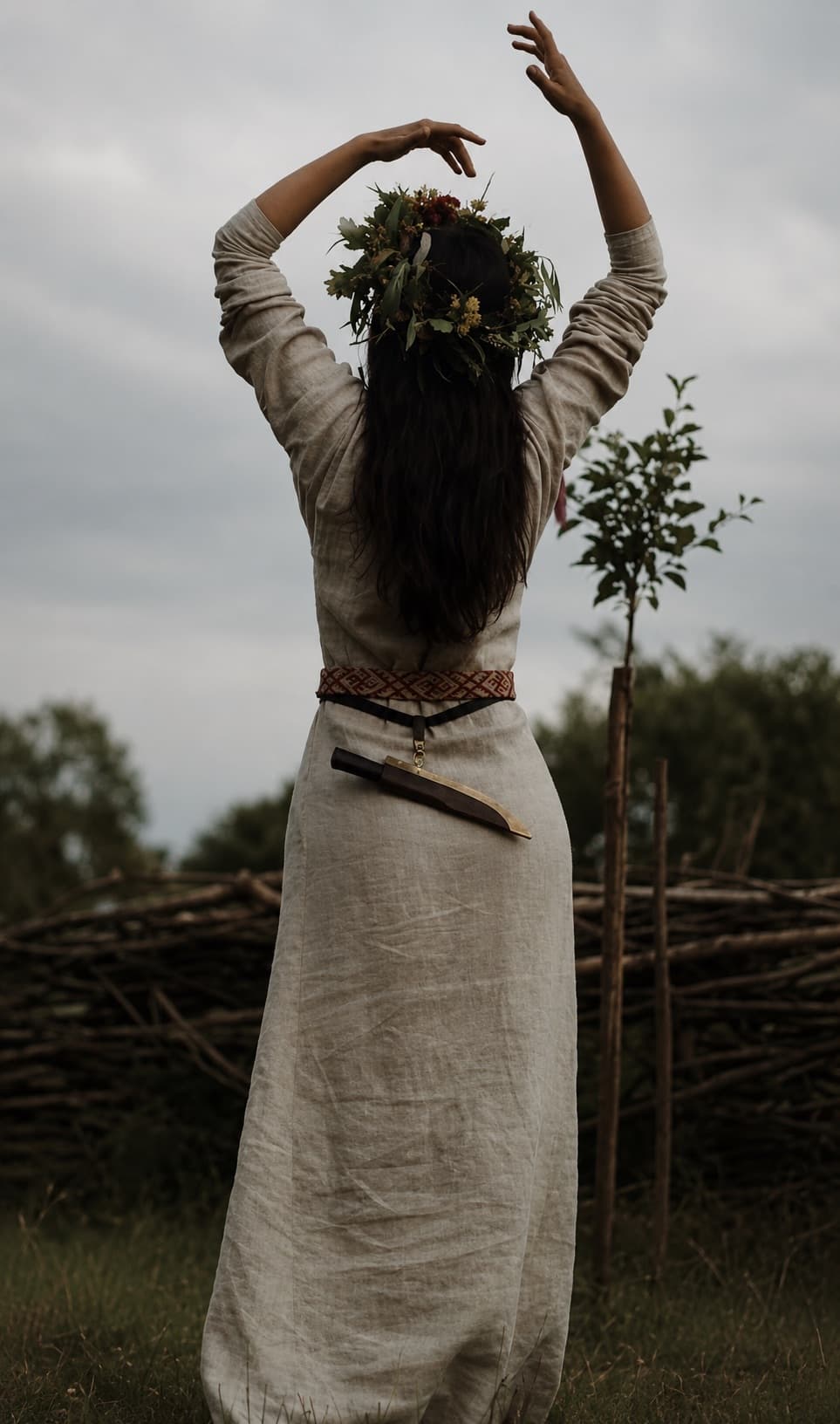 A woman in linen with a flower crown, arms raised to the sky in an open field